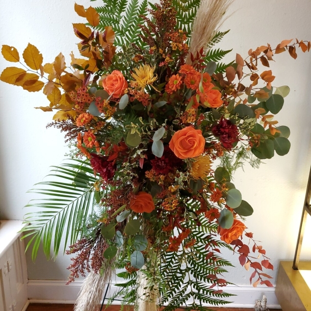 Copper Brook sympathy standing spray with orange roses, burgundy blooms, seasonal foliage, and greenery