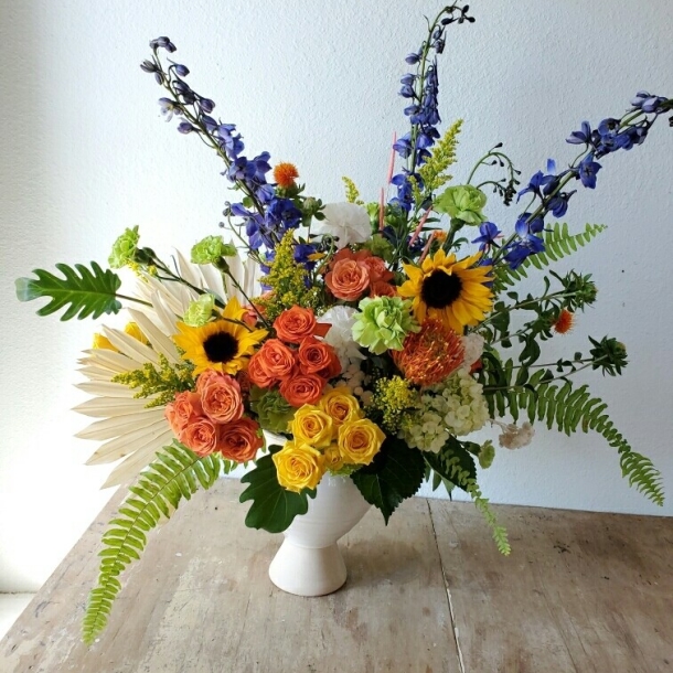 Janice floral arrangement with sunflowers, orange and yellow roses, blue delphinium, and lush greenery in a white vase