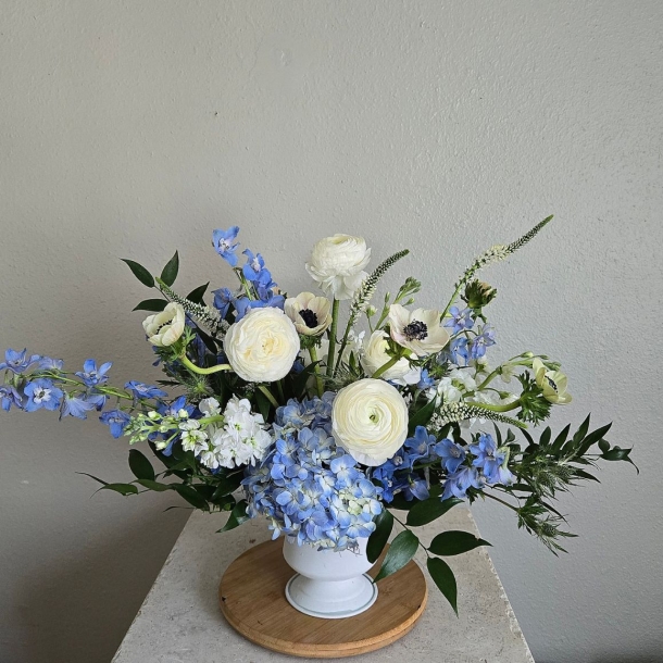 Naomi floral arrangement with blue hydrangeas, delphinium, and white ranunculus in a white pedestal vase