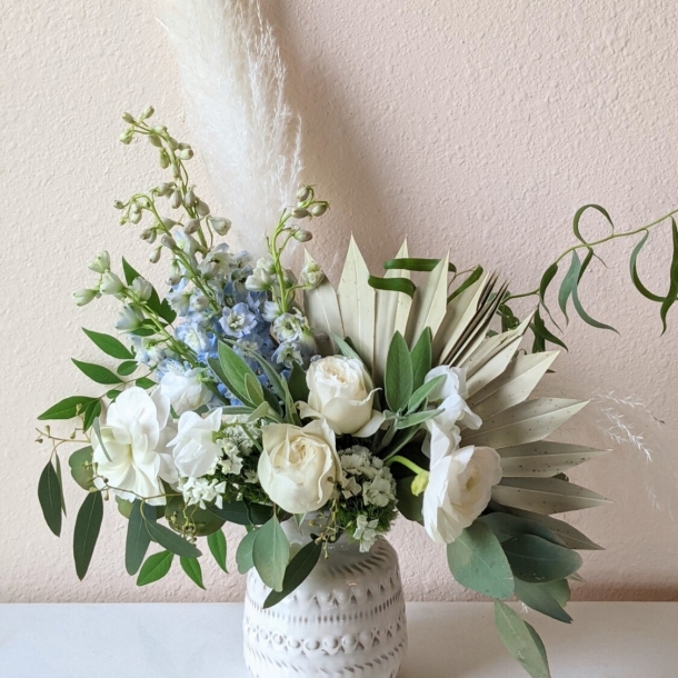 Thalia floral arrangement with white roses, pale blue blooms, eucalyptus, and dried palm leaves in a ceramic vase